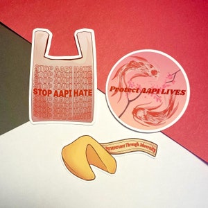 May include: Three stickers with messages of support for the AAPI community. The first sticker is a white plastic bag with the text "STOP AAPI HATE" repeated multiple times. The second sticker is a pink circle with two red fish and pink flowers, and the text "Protect AAPI LIVES". The third sticker is a yellow fortune cookie with the text "Perseverance Through Adversity".