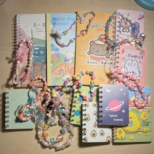 Puede incluir: Una colecci&oacute;n de peque&ntilde;os cuadernos coloridos con cadenas de cuentas. Los cuadernos tienen varios dise&ntilde;os, incluyendo un oso de dibujos animados, una escena espacial y un conejito lindo. El texto en los cuadernos incluye "Deep Forest Manor", "Bread is Okay", "Bake Bear", "Sweet", "Space Travel" y "Ollie's Life".