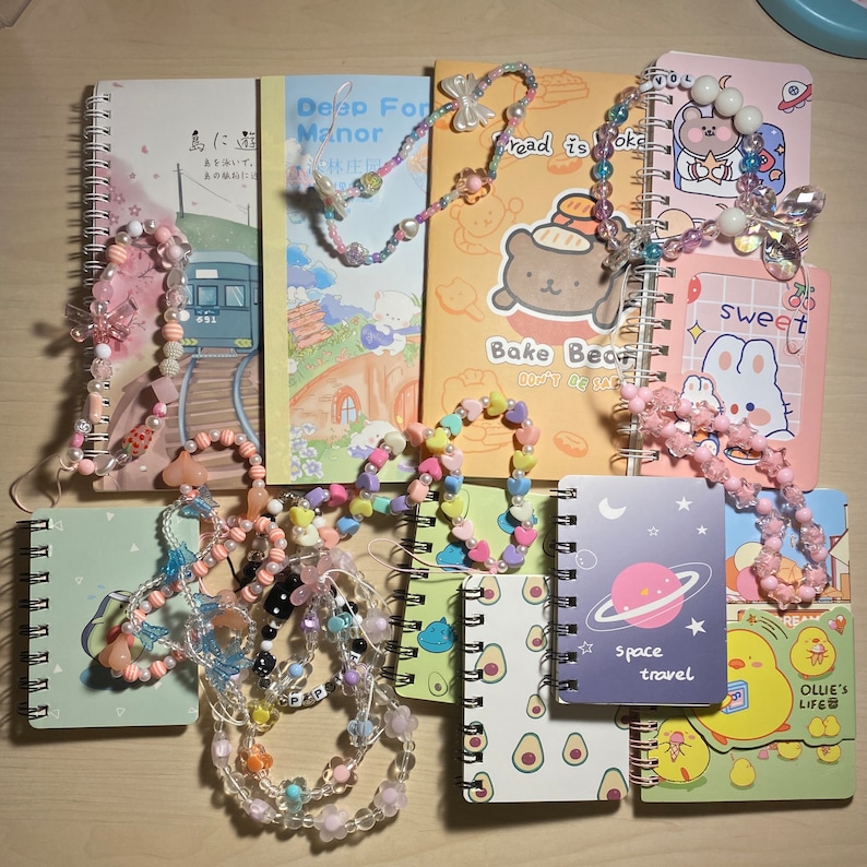 Puede incluir: Una colecci&oacute;n de peque&ntilde;os cuadernos coloridos con cadenas de cuentas. Los cuadernos tienen varios dise&ntilde;os, incluyendo un oso de dibujos animados, una escena espacial y un conejito lindo. El texto en los cuadernos incluye "Deep Forest Manor", "Bread is Okay", "Bake Bear", "Sweet", "Space Travel" y "Ollie's Life".