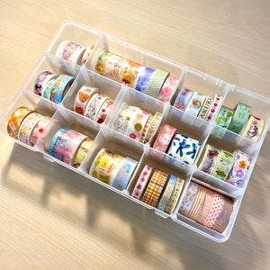 Puede incluir: Una caja de almacenamiento de pl&aacute;stico transparente llena de rollos de cinta washi de colores. La caja tiene 16 compartimentos y est&aacute; llena de varios patrones y colores de cinta washi.