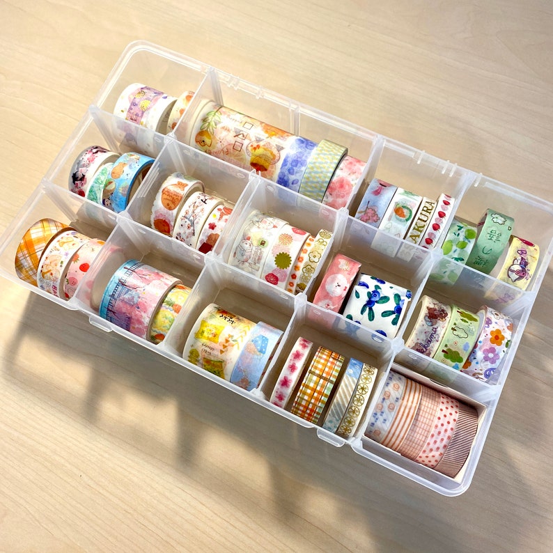 Puede incluir: Una caja de almacenamiento de pl&aacute;stico transparente llena de rollos de cinta washi de colores. La caja tiene 16 compartimentos y est&aacute; llena de varios patrones y colores de cinta washi.