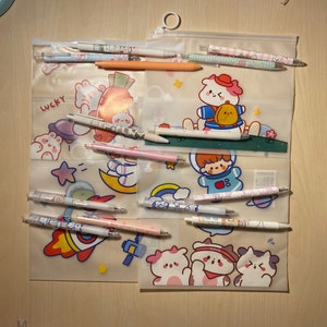 Puede incluir: Un estuche de l&aacute;pices de pl&aacute;stico transparente con cierre de cremallera. El estuche est&aacute; lleno de bol&iacute;grafos y l&aacute;pices de colores con lindos personajes de dibujos animados y dise&ntilde;os. El estuche tiene un fondo blanco con una variedad de personajes de dibujos animados, incluyendo un conejo, un oso y un gato. El texto "Lucky" est&aacute; impreso en el estuche.