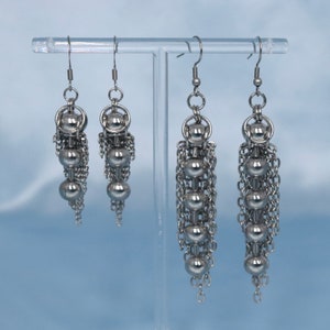 May include: Three pairs of silver dangle earrings with multiple small silver beads and chains. The earrings are displayed on a clear stand.