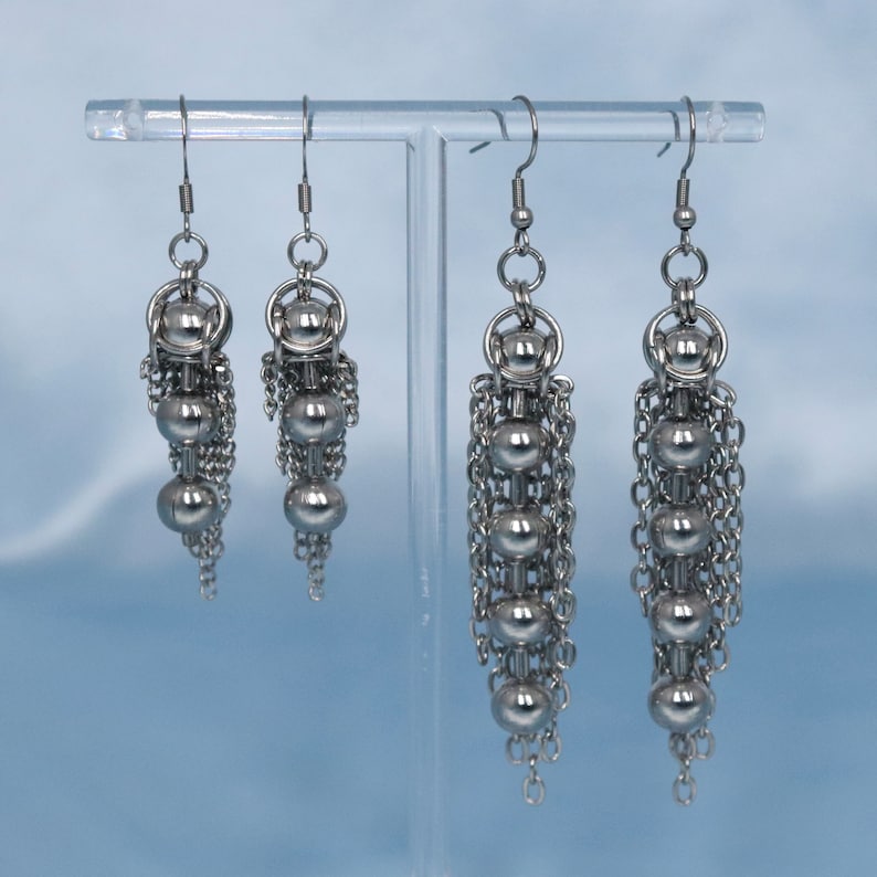 May include: Three pairs of silver dangle earrings with multiple small silver beads and chains. The earrings are displayed on a clear stand.