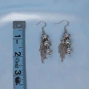 May include: Silver dangle earrings with a chain and two small ball charms. The earrings are approximately 5 cm long.