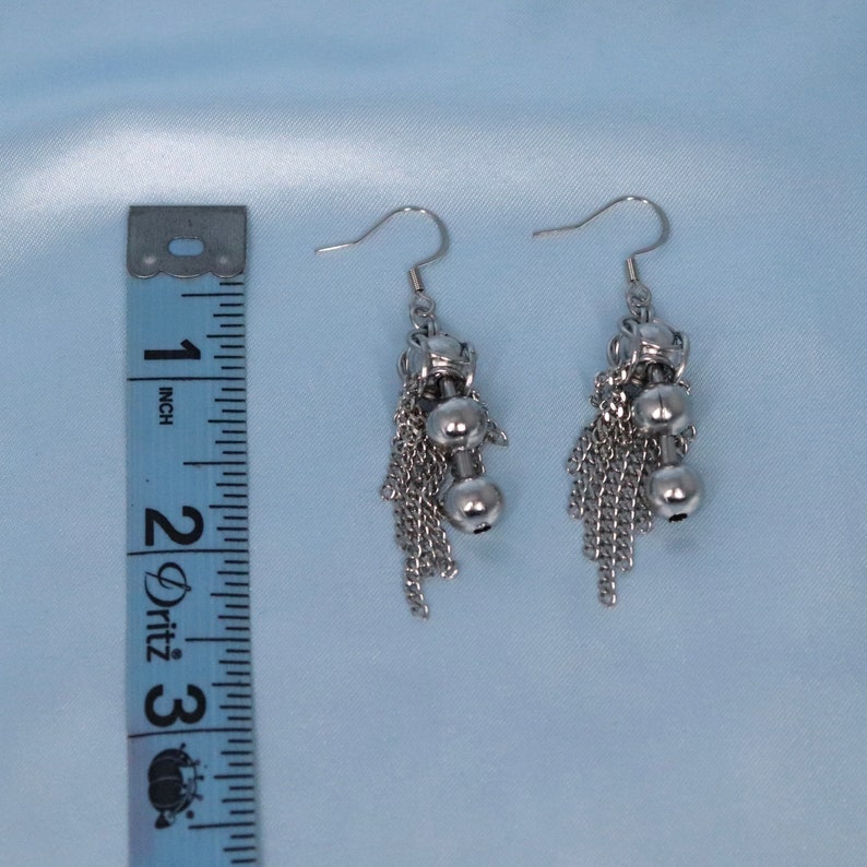 May include: Silver dangle earrings with a chain and two small ball charms. The earrings are approximately 5 cm long.