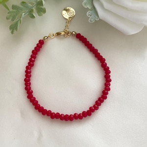 May include: A delicate red beaded bracelet with a gold-colored clasp and charm. The bracelet is made of small, faceted red beads strung together. The gold charm is round and has a small design. The bracelet is displayed on a white surface.