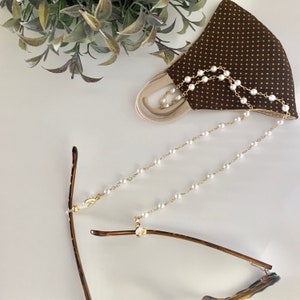 May include: A gold chain with white pearl beads, attached to a pair of brown glasses and a brown fabric face mask with white polka dots.