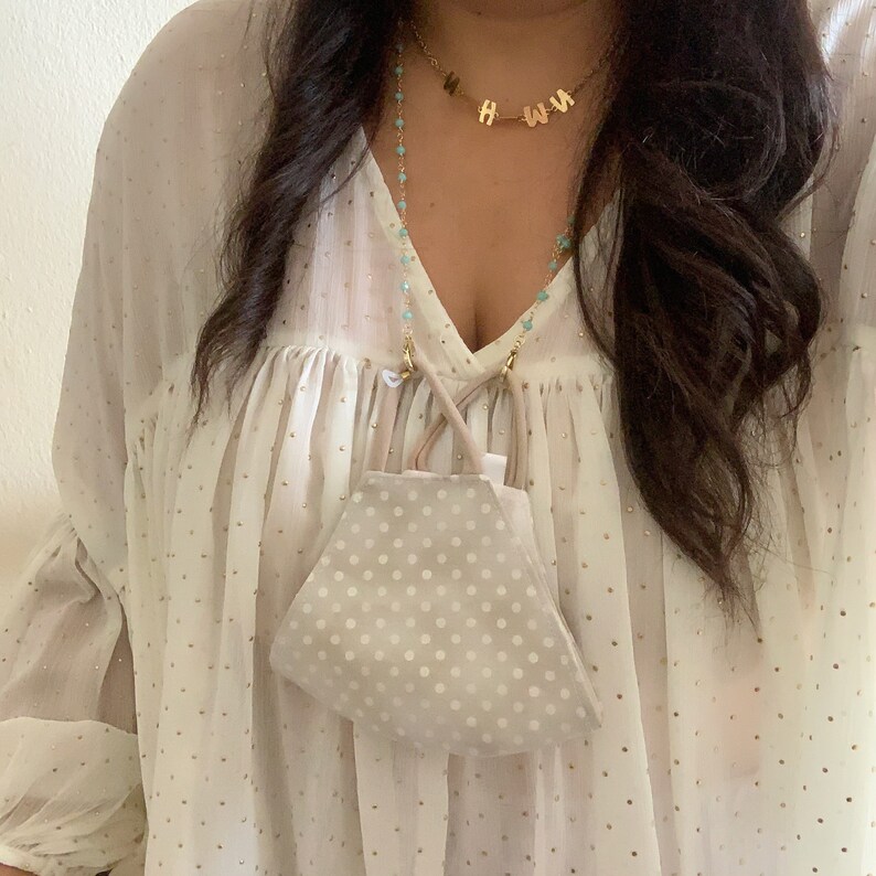 May include: A white blouse with gold polka dots and a turquoise beaded chain with gold clasps holding a beige polka dot face mask.