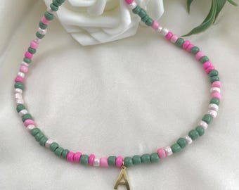 May include: A beaded necklace featuring a gold-colored letter "A" pendant. The necklace is composed of small, round beads in shades of pink, green, and white. The beads are strung together to form a delicate chain, suitable for everyday wear.