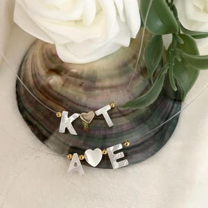 Puede incluir: Un delicado collar de plata con dos juegos de letras y corazones. El primer juego deletrea "KT" con un corazón dorado en el medio. El segundo juego deletrea "AVE" con un corazón blanco en el medio.