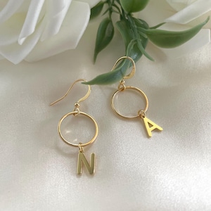 Puede incluir: Pendientes de aro dorados con las letras "A" y "N" colgando. Se muestran sobre una superficie blanca y sedosa, con una flor blanca y hojas verdes en el fondo.