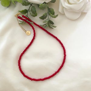 Puede incluir: Un delicado collar de cuentas rojas con un cierre dorado y un pequeño dije redondo. El collar se exhibe sobre una superficie blanca texturizada, con una rosa blanca y follaje verde en el fondo.