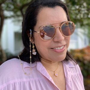 May include: A woman wearing round, brown sunglasses with a gold chain attached to the frames. The chain has small gold beads and pink and white charms. She is also wearing a gold necklace with a pink heart pendant.