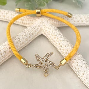 以下が含まれることがあります： クリアクリスタルをちりばめたゴールドのヒトデペンダントが付いた、黄色の調節可能なブレスレット。ブレスレットは、ゴールドトーンの金属アクセントと織りロープのデザインです。ヒトデのペンダントが中心で、海の魅力を添えています。