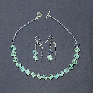 May include: A silver necklace and earrings set featuring green and blue fluorite gemstone beads. The necklace has a silver toggle clasp and the earrings have silver hooks.