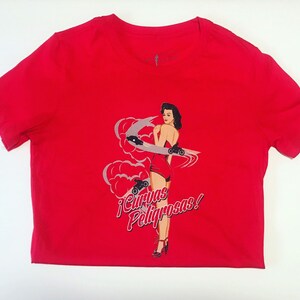 K&ouml;nnte beinhalten: Rotes T-Shirt mit einem Aufdruck einer Frau in einem roten Kleid und Rollschuhen. Der Text auf dem Shirt lautet "&iexcl;Curvas Peligrosas!"