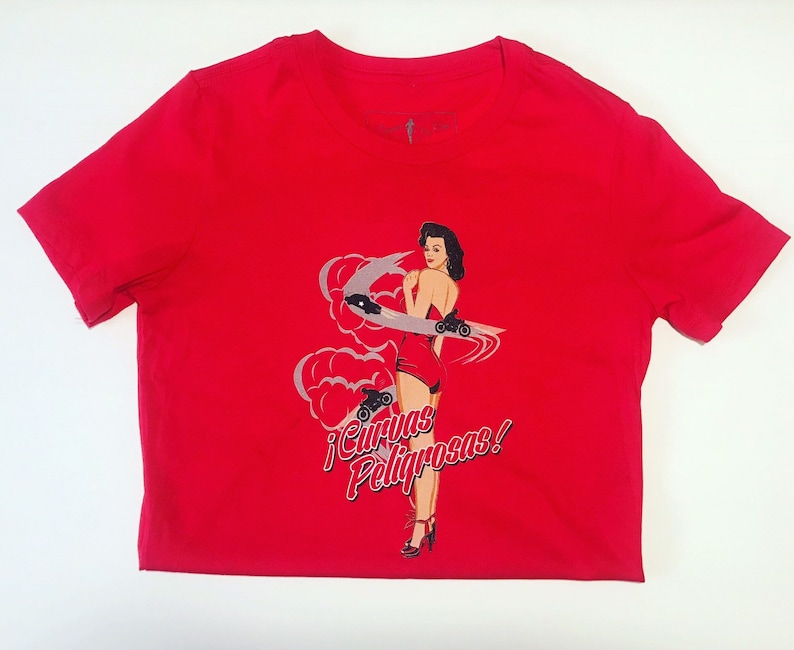 K&ouml;nnte beinhalten: Rotes T-Shirt mit einem Aufdruck einer Frau in einem roten Kleid und Rollschuhen. Der Text auf dem Shirt lautet "&iexcl;Curvas Peligrosas!"