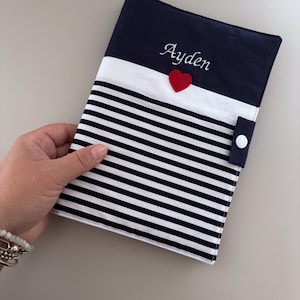 Puede incluir: Una funda de tela para cuaderno con una sección superior azul marino que presenta el nombre Ayden bordado en letra blanca, un pequeño corazón rojo aplicado y una sección inferior con rayas horizontales negras y blancas, asegurada por una lengüeta azul marino con un broche blanco.