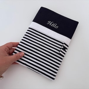 Peut inclure: Une pochette en tissu rayé bleu marine et blanc avec le nom "Hélio" brodé en blanc. La pochette a une bande blanche et une fermeture à bouton noire. La pochette est tenue par une main.