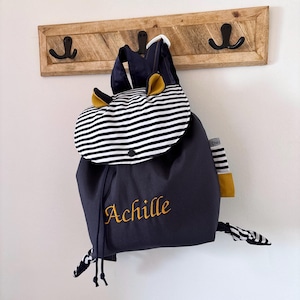 Sac à dos enfant en tissu bleu marine, suspendu à un porte-manteau en bois fixé au mur. Rabat à rayures marines et blanches avec bouton-pression, petites oreilles décoratives de couleur jaune. Prénom « Achille » brodé en jaune sur la face avant. Bretelles et cordons de serrage assortis. Sac à dos personnalisé pour enfant.