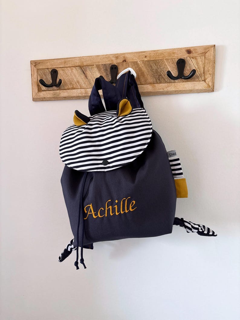 Sac à dos enfant en tissu bleu marine, suspendu à un porte-manteau en bois fixé au mur. Rabat à rayures marines et blanches avec bouton-pression, petites oreilles décoratives de couleur jaune. Prénom « Achille » brodé en jaune sur la face avant. Bretelles et cordons de serrage assortis. Sac à dos personnalisé pour enfant.