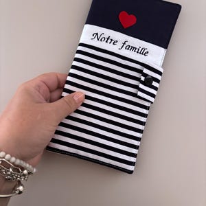 Può includere: Un portadocumenti rettangolare in tessuto con una sezione superiore blu navy decorata con una piccola applicazione a forma di cuore rosso, una fascia bianca con il testo ricamato in nero "Notre famille" e una parte inferiore a righe bianche e nere con chiusura a scatto.
