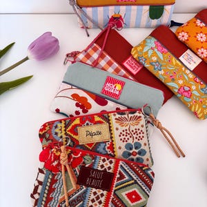 May include: A collection of colorful zippered pouches with various patterns and textures. Some pouches feature floral, geometric, and striped designs. Several pouches have labels with text like "YOU'RE THE ONE THAT I WANT" and "SALUT BEAUTÉ".