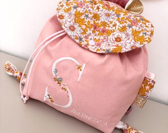 Puede incluir: Una mochila de cordón rosa con una solapa de estampado floral y orejas de purpurina dorada. El frente está bordado con una letra S blanca grande y el nombre haïneze k en letra cursiva blanca. La bolsa incluye cordones a juego con estampado.