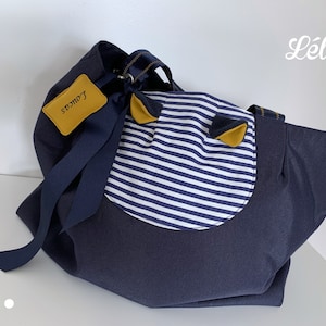 Peut inclure: Un sac fourre-tout en tissu bleu marine avec un empiècement circulaire rayé blanc et bleu marine sur le devant, deux petites oreilles jaune moutarde et bleu marine, et une étiquette jaune moutarde portant le nom Loucas en noir.