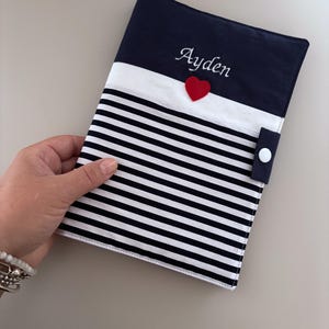 Peut inclure: Une couverture de livre à rayures bleu marine et blanches avec une bande blanche et un dessus bleu marine. Le nom "Ayden" est brodé en écriture blanche au-dessus d'un petit cœur rouge. Une languette bleu marine avec un bouton blanc sécurise la couverture.