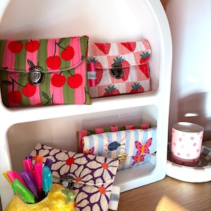 Puede incluir: Una estantería blanca exhibe varias bolsas de tela con diversos estampados, incluyendo cerezas, fresas, margaritas y mariposas. Las bolsas tienen cierres metálicos y el texto "Chez" en una pequeña etiqueta. Un portalápices de cerámica con forma de limón y rotuladores de colores está en primer plano.