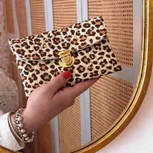 Könnte beinhalten: Eine gesteppte Clutch mit braun-schwarzem Leopardenmuster. Die rechteckige Tasche hat eine Umschlagklappe, die mit einem goldfarbenen, herzförmigen Druckknopfverschluss gesichert ist. Der Stoff hat eine gesteppte, texturierte und gepolsterte Optik.