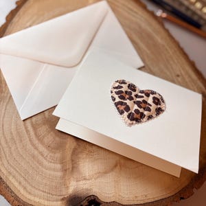 Peut inclure: Une carte de vœux pliée blanche ornée d'un cœur en tissu matelassé avec un motif imprimé léopard marron et noir. Le cœur est cousu sur le devant de la carte avec du fil visible. Une enveloppe blanche assortie est placée derrière la carte.