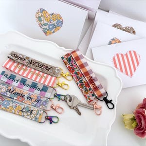 Puede incluir: Llaveros de tela con diseños florales y estampados, cada uno con texto en francés. Los llaveros se exhiben en una fuente blanca, junto con tarjetas blancas con adornos en forma de corazón y un ramo de rosas rosas.