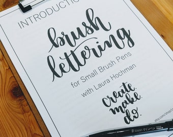 May include: A white sheet of paper with the text "INTRODUCTION TO brush lettering for Small Brush Pens with Laura Hochman Create make do" and a black brush pen on top of the paper.