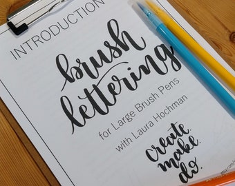 May include: A clipboard with a white sheet titled "Introduction to Brush Lettering" with black calligraphy. Three brush pens in blue, yellow, and orange rest on the sheet. The text "Create, make, do" is also visible.