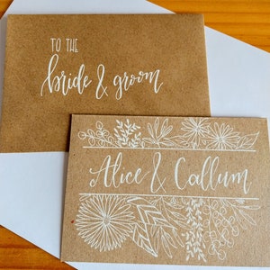 May include: A brown card with white calligraphy that reads "To the bride & groom" and a second card with white calligraphy that reads "Alice & Callum" and a floral design.