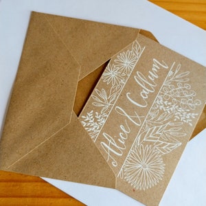 May include: A brown paper envelope with a white card inside. The card has a floral design and the text "Alice & Callum" written in white calligraphy.