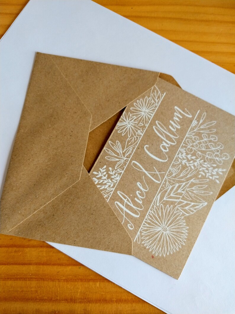 May include: A brown paper envelope with a white card inside. The card has a floral design and the text "Alice & Callum" written in white calligraphy.