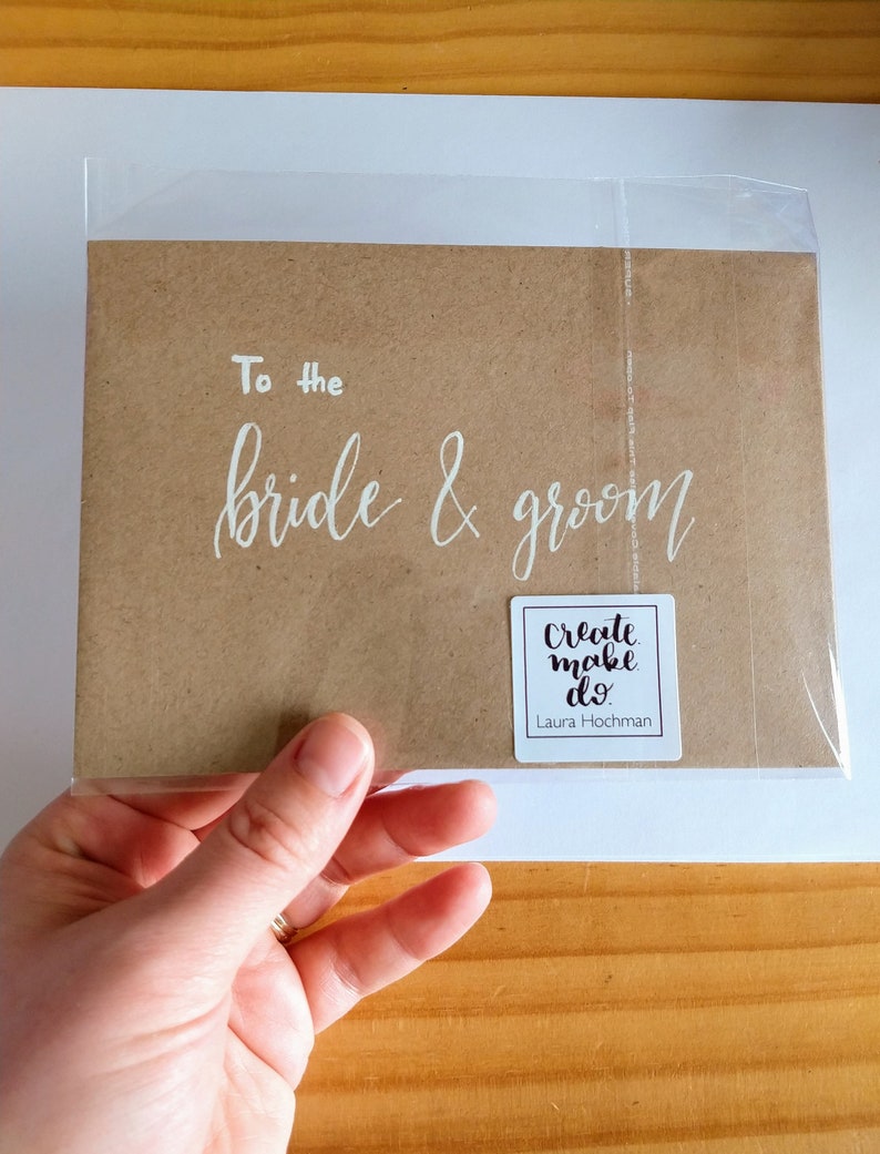 May include: A brown card with white calligraphy that reads "To the bride & groom". A white sticker with black text reads "Create make do" and "Laura Hochman".