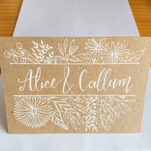 May include: A brown card with white ink calligraphy that reads "Alice & Callum" surrounded by a border of white floral designs.