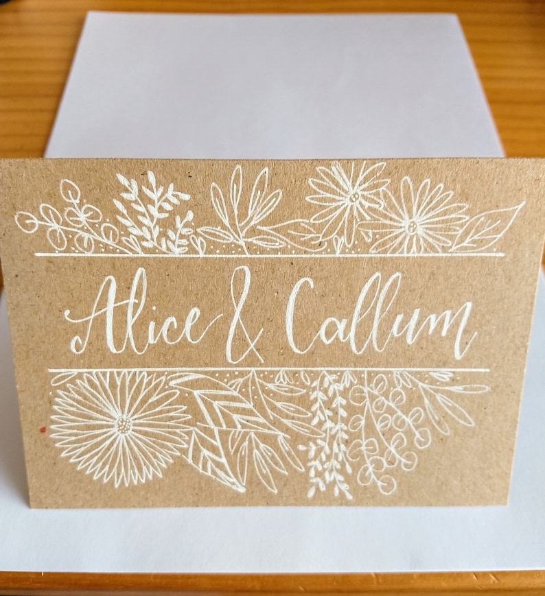 May include: A brown card with white ink calligraphy that reads "Alice & Callum" surrounded by a border of white floral designs.