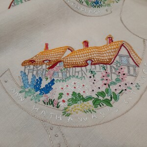 May include: Embroidered linen fabric with a cottage scene. The cottage has a thatched roof and is surrounded by flowers. The text "Ann Hathaway's Cottage" is embroidered around the scene.