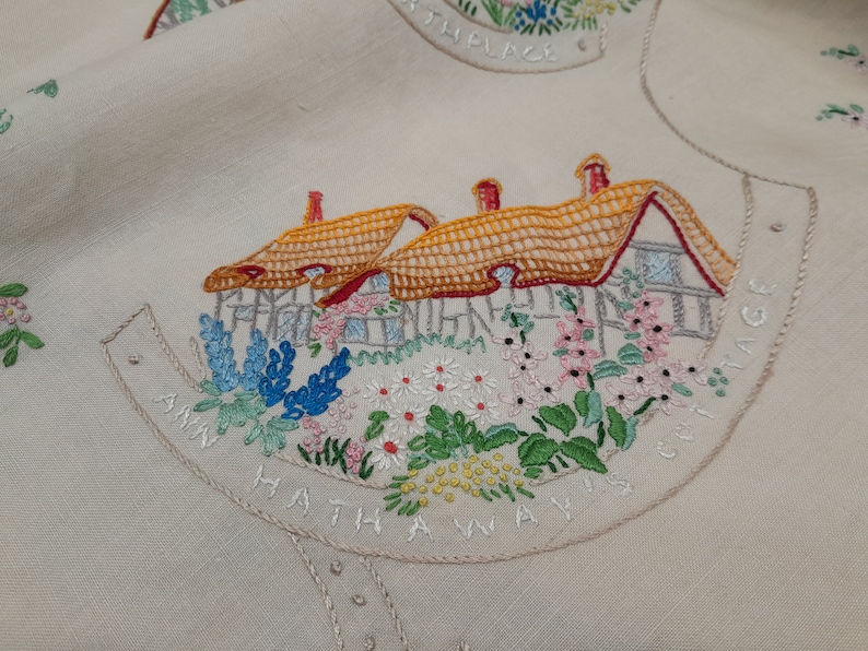 May include: Embroidered linen fabric with a cottage scene. The cottage has a thatched roof and is surrounded by flowers. The text "Ann Hathaway's Cottage" is embroidered around the scene.