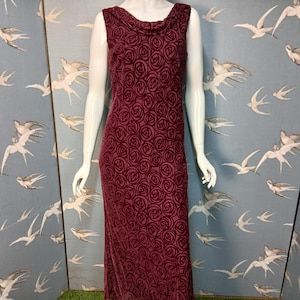 May include: Burgundy sleeveless dress with a cowl neck and a rose pattern. The floor-length dress is made of a velvet-like material. The dress is displayed on a mannequin.