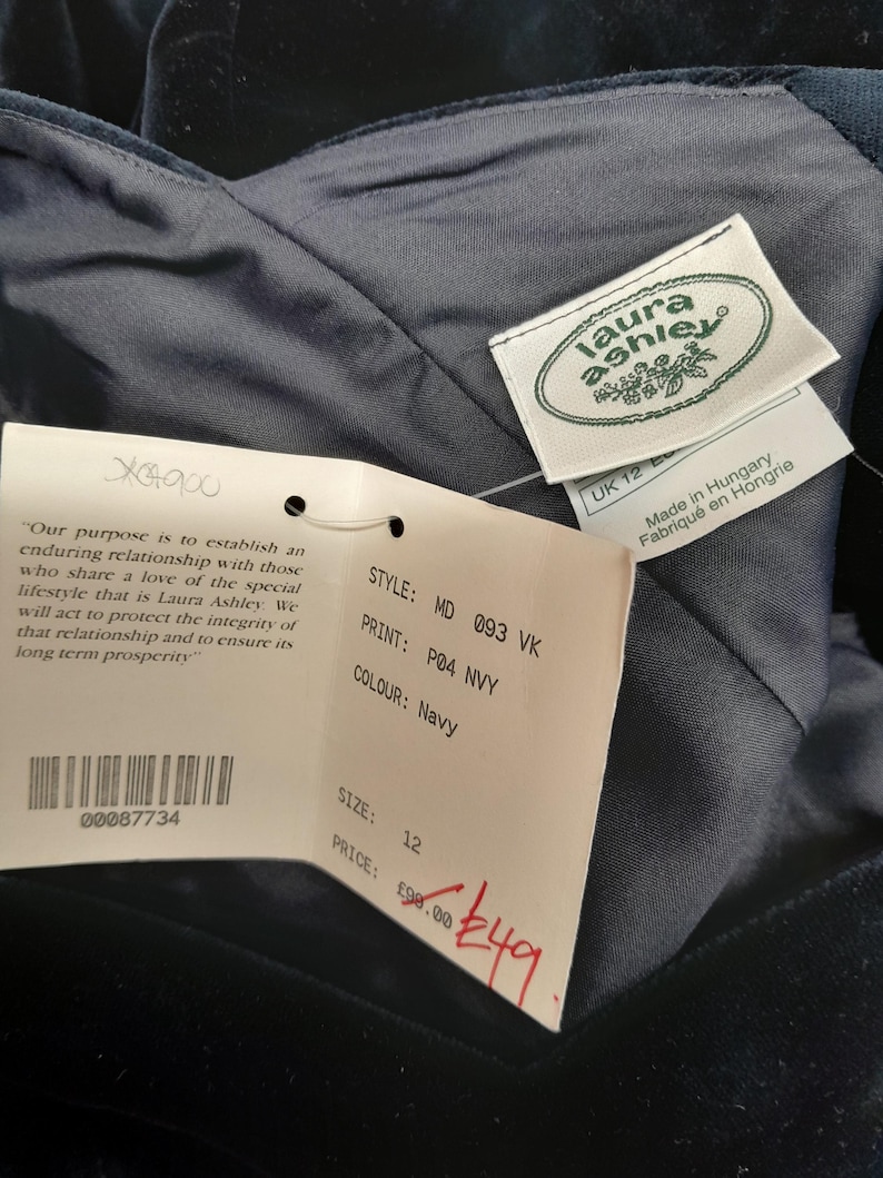 May include: A navy blue velvet dress with a Laura Ashley tag. The tag has the text "Laura Ashley" in a white font with a green leaf design. The tag also has the text "Made in Hungary" and "Fabriqu&eacute; en Hongrie". The dress has a price tag attached with the text "STYLE: MD 093 VK", "PRINT: P04 NVY", "COLOUR: Navy", "SIZE: 12", "PRICE: &pound;98.00", and "&pound;49".