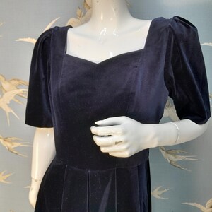 May include: A navy blue velvet dress with a square neckline and short puffed sleeves. The dress has a fitted bodice and a full skirt.