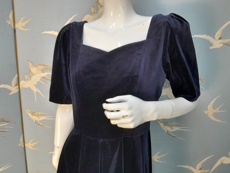 May include: A navy blue velvet dress with a square neckline and short puffed sleeves. The dress has a fitted bodice and a full skirt.