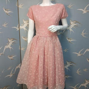 May include: A vintage pink dress with a floral pattern. The dress features a fitted bodice, short sleeves, and a flared skirt. The dress is displayed on a mannequin. The background is a light blue wall with a bird pattern.